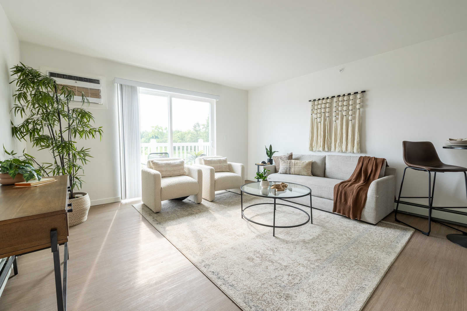 Bright living room with a cream sofa, two armchairs and a glass coffee table on a patterned rug, with a sliding glass door and a wall-mounted air conditioner